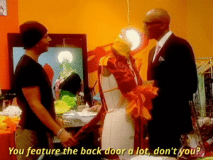 Two people talking with a mannequin dressform in between them, one wearing a black t-shit and beanie, the other wearing a suit, with the caption, “You feature the back door a lot, don’t you?”