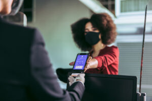 Female traveler wearing face mask showing electronic boarding pass on phone to ground attendant at check-in counter at airport terminal during pandemic.