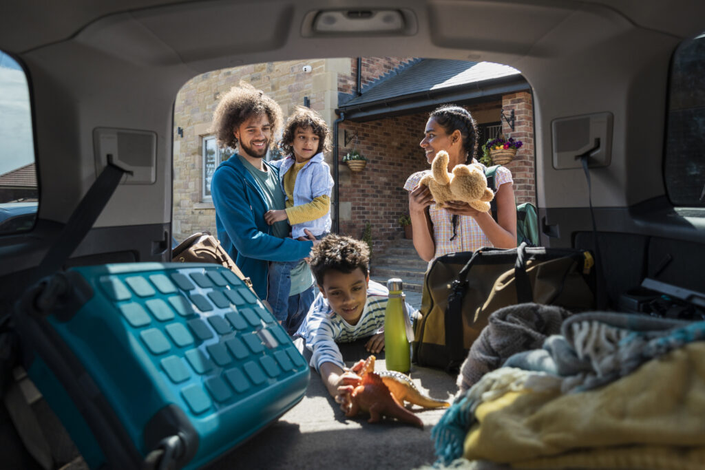 Young family packing their car trunk for a road trip as son reaches for a toy