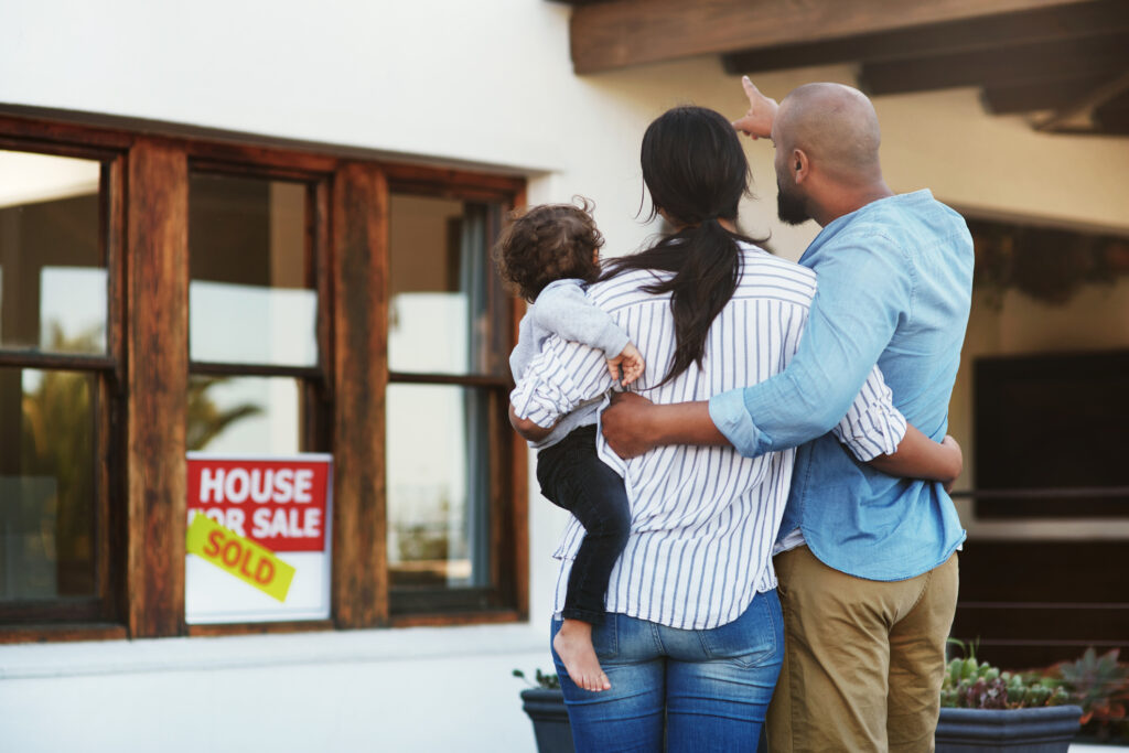 Shot of a young family of three facing their new home outside with a sold sign in the window file the father points towards the home